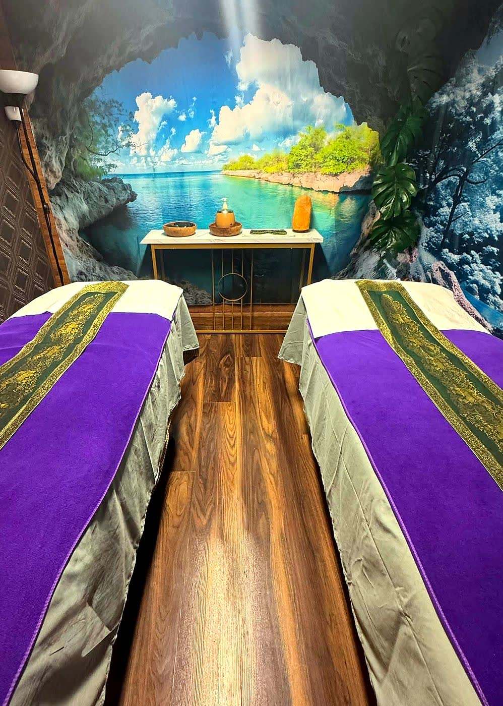The couples treatment room at Taitam-D: two massage beds dressed in purple with Thai gold brocade runners, against a painted lagoon mural